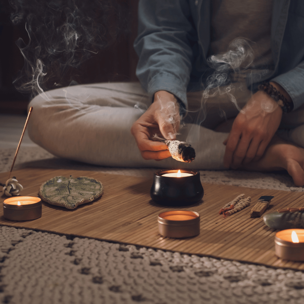 Rituale di purificazione: mani che agitano un fascio di salvia fumante sopra un bruciatore, con candele accese e bastoncini d’incenso su un piano in bamboo.